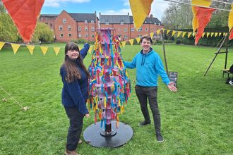 A woman and man stand either side of a large ornamental item in a field of grass with bunting hung up 