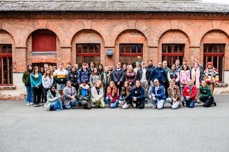 A large group of young people gathered together outside a red brick building with archway windows