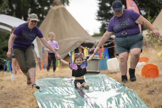 Two women hold a little girls hands as she slides down a water slide in an outdoor setting. The women's tshirts read Wildchild Festival