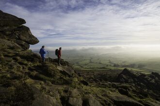 Two men stand overlooking a vast landscape from high on a hill with rocks behind them