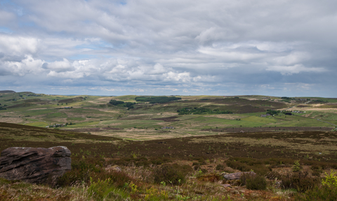 A vast moorland view over a patchworked landscape of farm fields with heather and rocks in the foreground