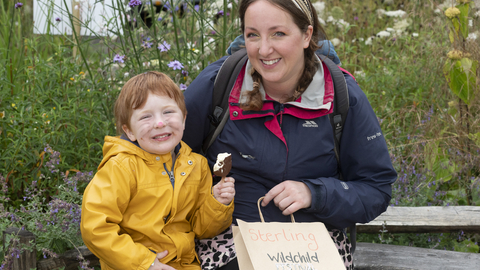 A woman and child sit in a wildflower garden smiling, the woman holds a wildchild paper bag and wears a head scarf, the child holds an icecream