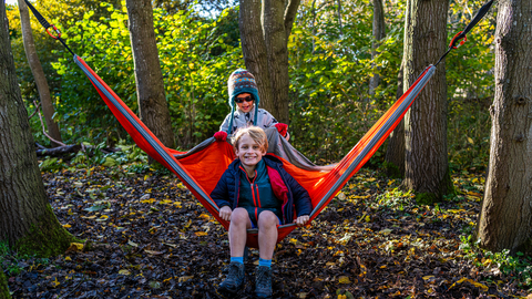 A boy with blonde hair sits in a hammock smiling with another boy in a woolley hat behind him