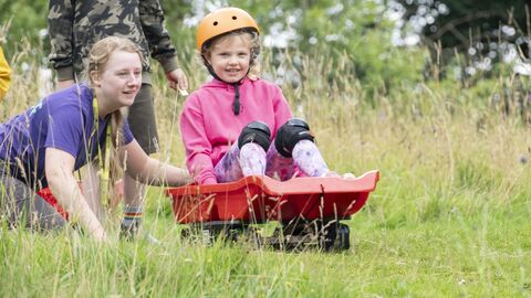 A girl sits on a red grass sled and looks excited as she rolls down a hill, a young woman in purple pushes her