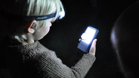 A young boy looks at a bat detector under the light of a headlamp.