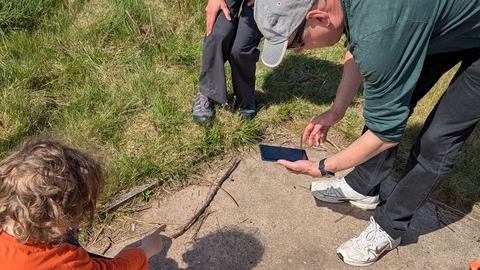 A woman stands watching as a man uses his phone to take a photo of something on the floor that a small child points at