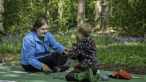A woman sits on a green tarpaulin on the woodland floor as she interacts with a young child