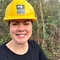 A woman smiling at the camera, wearing a black top and bright yellow hard hat