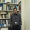 A man with dark hair and a moustache stands in front of a bookcase full of books, he wears a dark grey shirt and black trousers and stands next to a colourful canvas of accorns