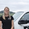 A woman with long blonde hair past her shoulders stands next to a white pick up truck and smiles. A large hill is behind her.