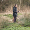 A woman stands next to a marshy pond area with a trail cam in her hands. She wears a black waterproof coat and has long dark hair which is tied up