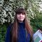A young woman with long mid brown hair with a fringe stands in front of a flowering blackthorn holding a clipboard. She wears a blue fleece.