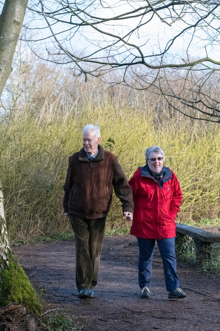 A man an woman both with grey hair walk hand in hand along a pathway surrounded by greenery and a tree