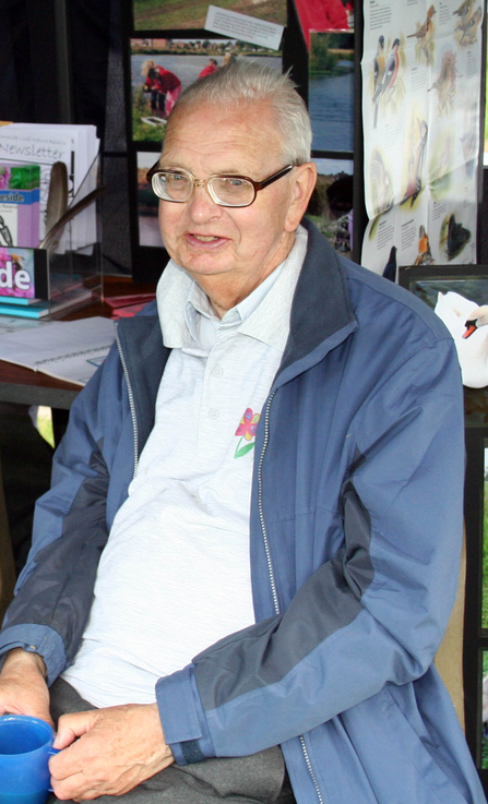 An elderly man sits next to a table holding a mug. He wears glasses, a white rugby style top and a blue waterproof coat