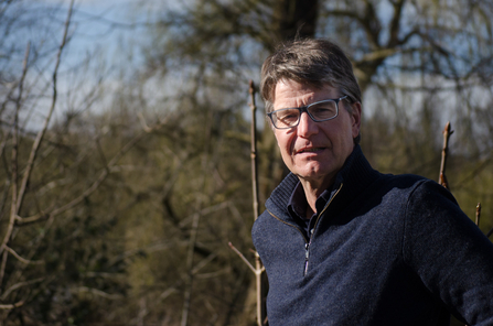 A man with dark hair and glasses, wearing a navy top stands with trees and bushes behind him