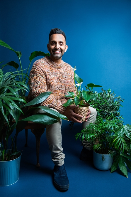 A man with black hair smiles at the camera as he sits amongst lots of green leafy plants in a studio with a plain blue background