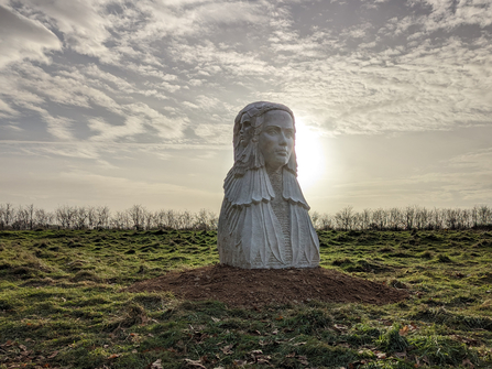 A large stone statue of a women with three faces stands on grass with trees in the distance, the sun is behind the statue with wispy clouds