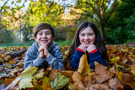 A brown haired boy and girl lie among the autumnal leaves on the floor. They prop their chins up with their hands and smile at the viewer. The boy wears a grey camo hoody and the girl wears a navy and red top.