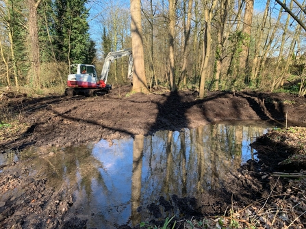 A digger near a freshly excavated pond surrounded by trees and bare earth.