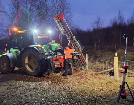 A tractor working in the dark to install new fencing, a man stands infront of the tractor in a high vis orange coat working