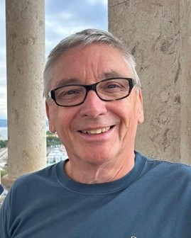A man smiles at the camera. He has short cropped grey hair and wears black rimmed glasses and a blue top.