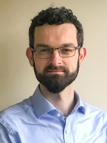 A middle aged man with short dark curly hair and a beard. He wears glasses and a pale blue shirt with the collar worn open.
