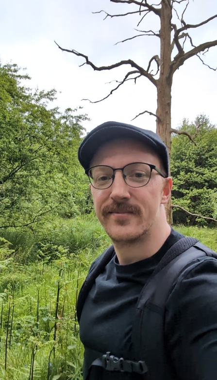 A man stands in front of a bare dead tree. He wears a baseball cap and glasses and has a moustache. He wears a dark top and backpack.