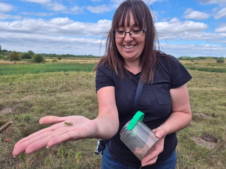 A woman with long brown hair and glasses stands in a rural area holding out her hand with a tiny grasshopper sitting on it. She smiles and looks very happy. She holds a bug pot and wears a black top.