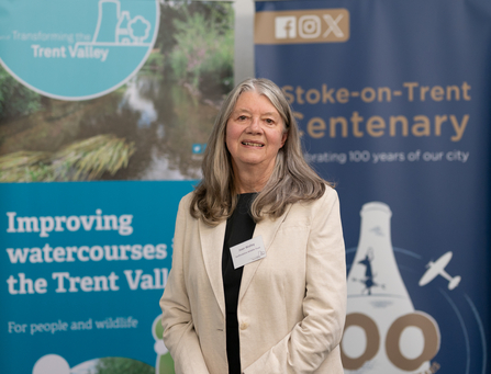 A woman with shoulder length dark grey hair smiles at the camera. She wears a beige jacket and black top and stands in front of two blue banners with text reading "improving watercourses the Trent Valley and Stoke-on-Trent Centenary