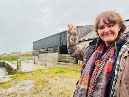 A woman stands in front of farm buildings holding a set of keys smiling