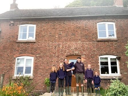 A family of 7 stand in front of a brick farmhouse lined up in order of height with the two adults in the middle and two children either side. The woman also holds a baby.