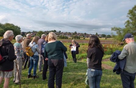 A group of people outside stand around a woman who holds a large informational board.