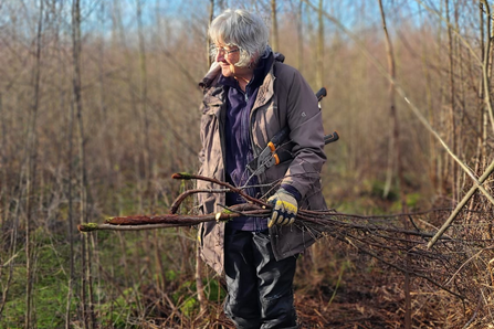 A woman wearing a jacket and gloves, standing amongst some trees with branches in her hand.
