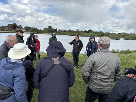 A group of people stand outside near a large pond on a wetland near an urban neighbourhood. 