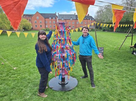 A woman and man stand either side of a large ornamental item in a field of grass with bunting hung up 