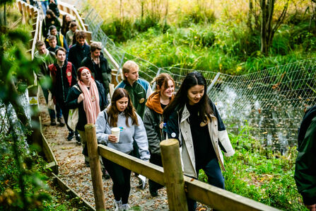 A large group of young people walking along a walkway in a wetland area surrounded by greenery