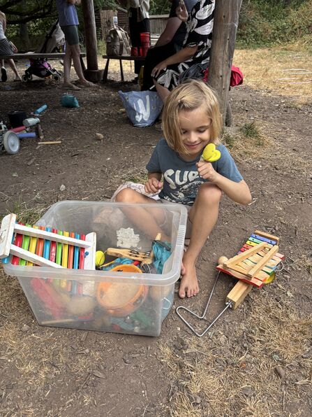 A young boy with chin length blond hair sits on the earth barefoot with a box of musical toys and smiles with joy