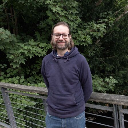 A man in a blue hoodie stands on a bridge with greenery behind him