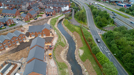 An aerial image shows a river meanders through an urban area with houses on the left and roads on the right