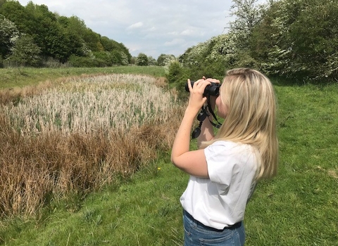 A teenage girl is looking for birds through binoculars, over a field surrounded by trees