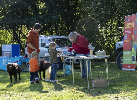 A woman reaches over a table set up outside to assist a child with making a creature out of clay. The family watches as the child makes art. Trees and gardening signs make up the background.