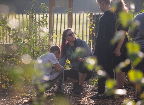 A women crouched down in a garden area with children surrounding her and a picket fence in the background