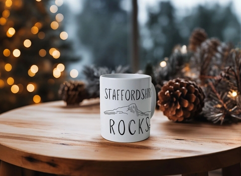 A white ceramic mug with Staffordshire Rocks text and the outline of a rocky landscape, sitting on a round wooden table with cones and Christ