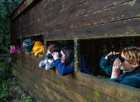 A group of children in brightly coloured clothing look out of a bird hide with binoculars 