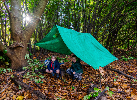 A bright green tarpaulin hangs over a line creating a den in a wooded area. Two boys crouch underneath the den roof and smile. A sunburt shines through the trees and lots of brown leaves cover the floor.