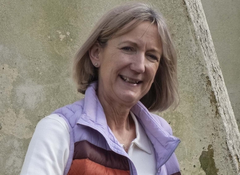 A woman smiles at the viewer, she has chin length cropped dark blonde hair and wears a white long sleeve tshirt with a lilac gilet that has brown and orange stripes.
