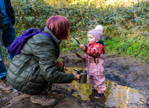 A woman bends down holding a metal tea pot as a small child holds a muddy spoon up to her face as she stands in a large muddy puddle in pink waterproofs