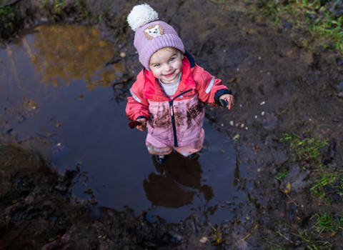 A young girl toddler stands in a muddy puddle - she wears a pink waterproof onsie suit and woolen bobble hat