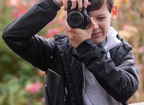 A teenage boy with short brown holds a camera to his face to take a photo. He wears a grey hoodie and black waterproof coat..