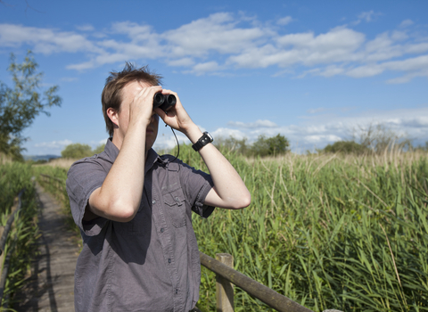 A man in a short sleeved top stands on a boardwalk in the sunshine with binoculars held up to his face.